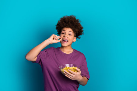 Smiling teenage boy enjoying a snack from a bowl against a vibrant blue background, expressing happiness and casual styleの写真素材