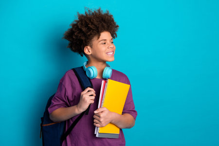 Smiling boy with curly hair holding notebooks against bright blue background, embodying trendy and cheerful school lifestyleの写真素材
