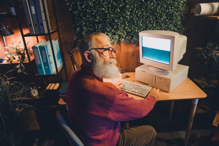 Elderly man working on vintage computer in a cozy home officeの写真素材