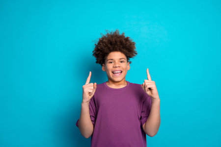 Happy young boy gesturing with fingers up against a bright blue backdrop wearing a casual purple...