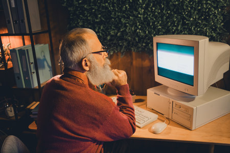 Senior man working thoughtfully on a vintage computer in a cozy home office during the evening