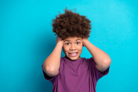 Smiling boy with hands on head against vibrant blue background showing surprise and joy wearing a casual purple t-shirtの写真素材