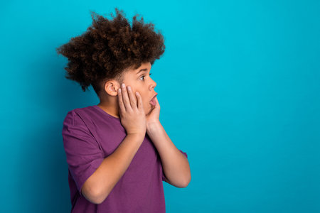 Excited young boy with curly hair expressing surprise and amazement against blue background in...