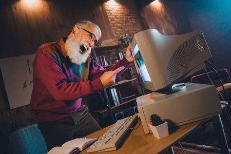 Elderly man enthusiastically engaging with vintage computer technology in a cozy home studio setting during evening hoursの写真素材