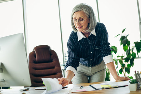 Confident mature businesswoman analyzing documents in modern office environment, showcasing professionalism and expertiseの写真素材