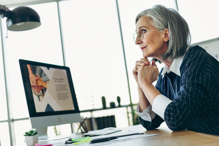Confident mature businesswoman with gray hair working in a professional office environment using a desktop computerの写真素材