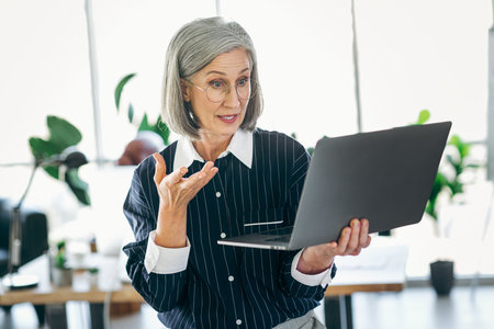 Confident mature businesswoman in formal attire standing in modern office, engaging in a video conference or online meeting.の写真素材