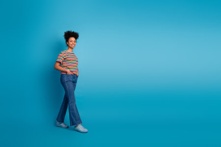 Confident young woman with afro hairstyle in colorful outfit standing against bright turquoise studio backgroundの写真素材