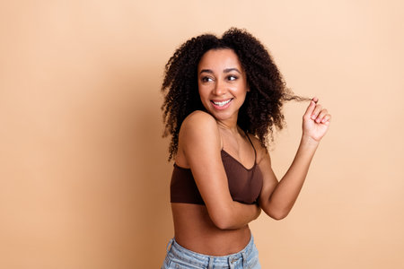 Young woman with curly hair in casual attire posing happily against a beige background, showcasing natural beauty and styleの写真素材