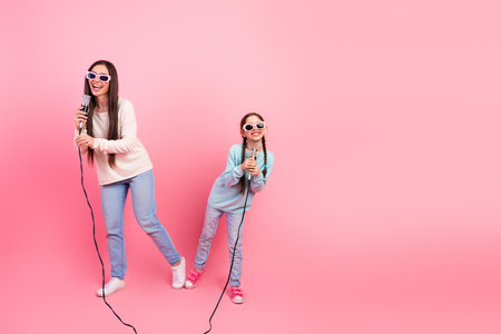 Happy mother and daughter singing karaoke together with colorful microphones against a vibrant pink wall in casual outfitsの写真素材