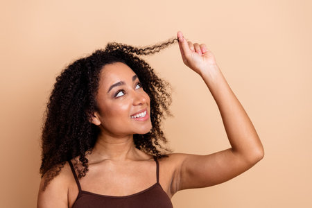 Portrait of a young woman with curly hair holding a strand, smiling, posing in stylish casual wear, against beige background.の写真素材