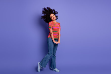 Joyful young woman with curly hair in vibrant casual outfit standing on purple background,...