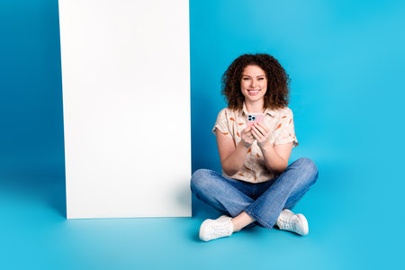 Young Woman With Curly Hair Smiling While Seated on Blue Background Holding a Smartphone in Casual...