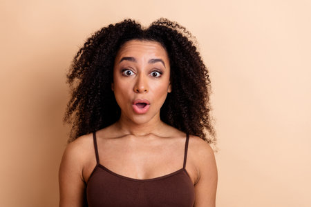 Young woman with curly hair expresses surprise while wearing a brown top against a beige background, showcasing emotionsの写真素材