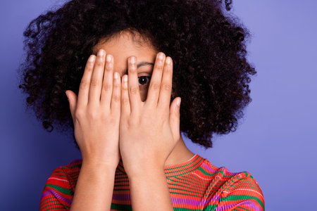 Youthful girl posing playfully with hands covering face against vibrant purple backdrop in cheerful...