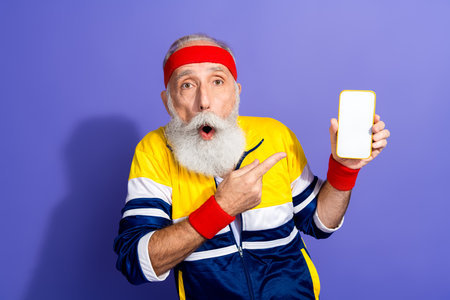 Elderly man wearing retro sportswear showcasing a smartphone, expressing surprise and humor against...