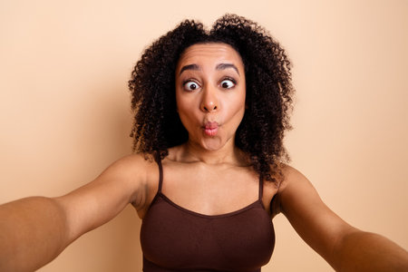 Playful young woman making a funny fish face and taking a cheerful selfie against a beige background