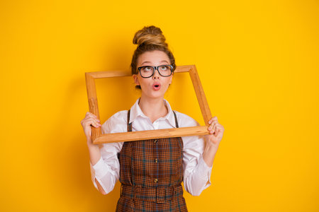 Cheerful young student holds a wooden frame, expressing surprise and joy, against a vibrant yellow backgroundの写真素材