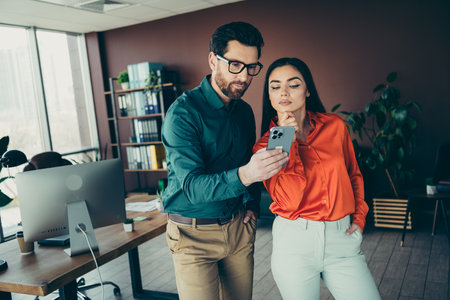 Two professionals discussing business tasks on a smartphone in a modern office setting with natural lightingの写真素材