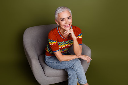 Smiling mature woman sitting in a modern chair wearing a colorful striped top and blue jeans against...