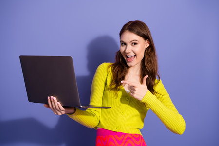 Young woman in stylish cardigan presenting laptop with excitement and charm against violet backgroundの写真素材