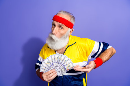 Fit elderly man in retro sportswear flaunting cash in a humorous pose against purple background