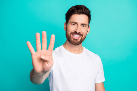 Smiling young man showing four fingers on turquoise background gesturing with hand illustrating...