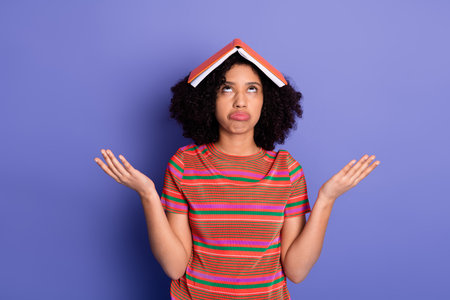 Young woman with book on her head making a frustrated gesture in colorful outfit on vibrant purple backgroundの写真素材