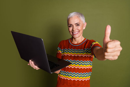 Smiling mature woman with short grey hair holding a laptop and giving a thumbs up on a khaki backgroundの写真素材