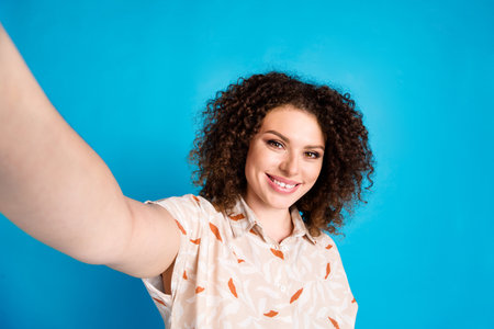 Smiling young woman taking a selfie against a vibrant blue wall, expressing happiness, casual style,...