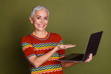 Happy senior woman with short grey hair using laptop on khaki background, showcasing a stylish...