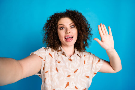 Cheerful young woman waving hand in a casual shirt against a vivid blue background expressing friendly and joyful gesturesの写真素材