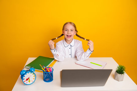 Cheerful young schoolgirl in white uniform studying at desk with laptop and book on bright yellow backgroundの写真素材