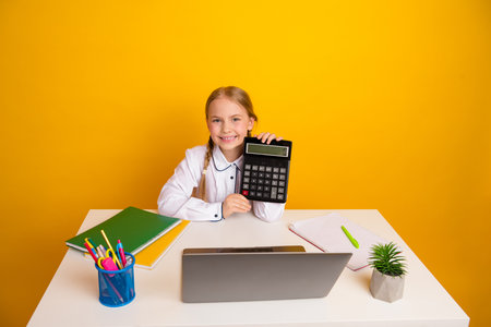 Elementary school girl holds a calculator while studying, sitting at a desk with supplies against a yellow backgroundの写真素材