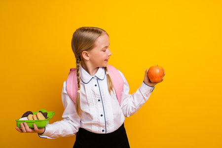 Happy schoolgirl chooses healthy snacks holding apple and lunchbox against a vibrant yellow backgroundの写真素材