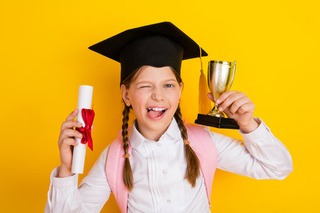 Cheerful schoolgirl posing with graduation cap, trophy, and diploma, standing against yellow backdrop celebrating achievementの写真素材