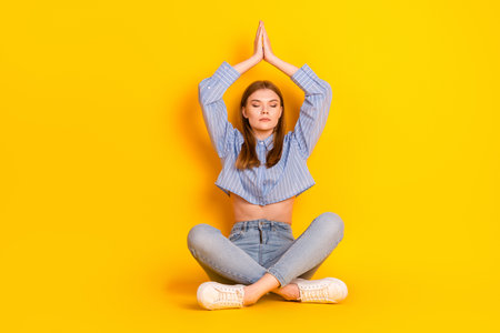 Young woman practicing mindfulness meditation on yellow background wearing casual blue shirt and jeans, serene and composedの写真素材