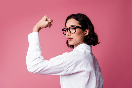 Charming young woman posing in business attire with modern eyewear against a bright pink backgroundの写真素材
