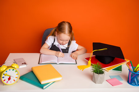 Little schoolgirl reading book at desk with colorful notebooks and academic hat in classroom setting