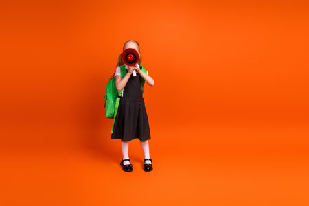 Young schoolgirl with red hair in uniform holding a megaphone on orange background expressing energy and enthusiasmの写真素材