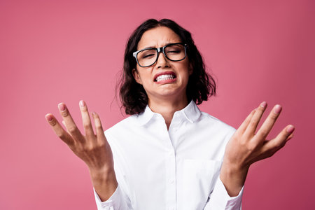 Young woman with expressive face and outstretched hands against pink background wearing formal white...