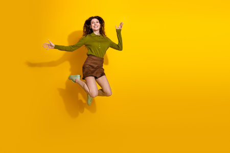 Cheerful young woman jumping with joy against a vivid yellow background, expressing happiness and...