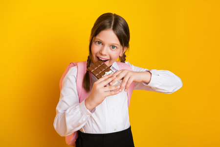 Smiling schoolgirl holding chocolate bar in hands standing against vibrant yellow background wearing uniform with braidsの写真素材