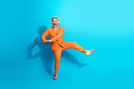 Charismatic mature man in vibrant orange suit dancing joyfully on bright blue background, portraying happinessの写真素材