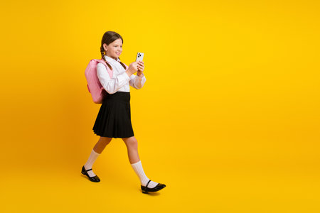 Happy schoolgirl in a uniform with backpack using smartphone on a yellow backgroundの写真素材