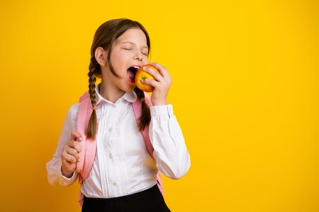 Adorable schoolgirl eating an apple on a bright yellow background, dressed in a white shirt with a backpack, cheerful and happyの写真素材