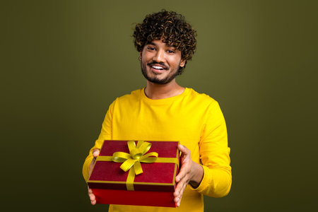 Happy young man with curly hair holding a gift box and smiling on a khaki background in casual yellow attireの写真素材