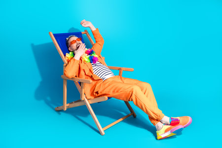 Vibrant man in an orange outfit relaxing in a deck chair against a bright blue backdrop during a festive celebrationの写真素材