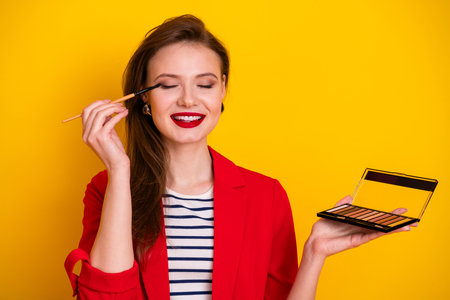 Young woman applying makeup with eyeshadow palette against a vibrant yellow backgroundの写真素材