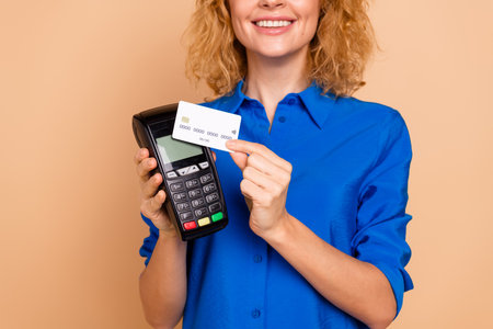 Smiling woman showcasing payment with a card on a portable terminal while wearing a stylish blue shirt against a beige backgroundの写真素材